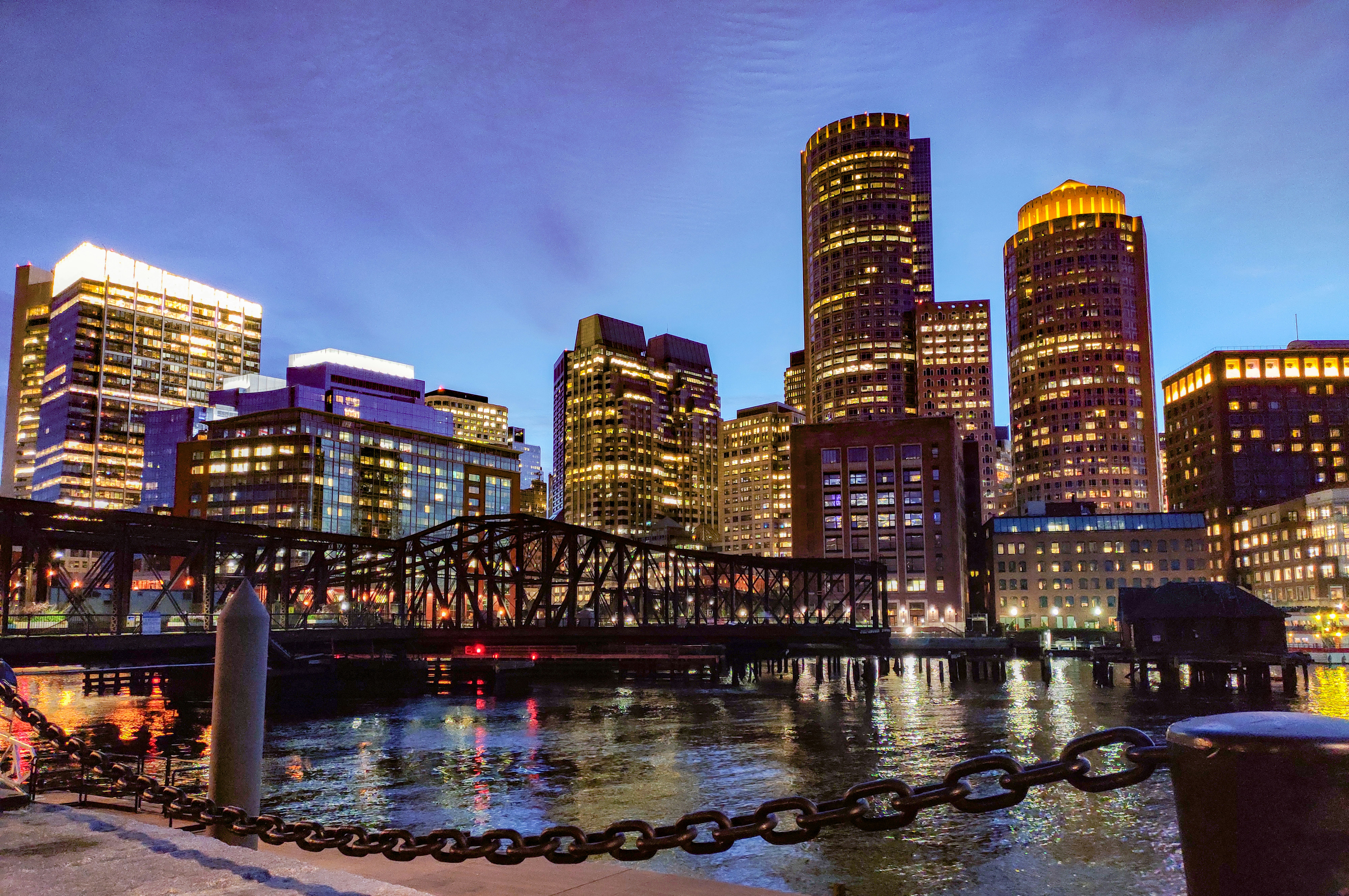 Image of downtown Boston, MA in the evening. Image includes some water with a bridge, buildings, and a darkened sky in the background. 